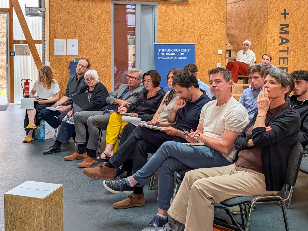 Männer und Frauen widmen sich sitzend interessiret den Vorträgen zu de Abschlussarbeiten der Architekturstudierenden der TU Darmstadt.