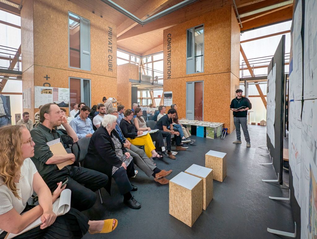 Unterschiedlichste Menschen sind zu Gast bei der Abschlusspräsenation der Architketurstudierenden und schauen interessiert zur gerade vortragenden Person.